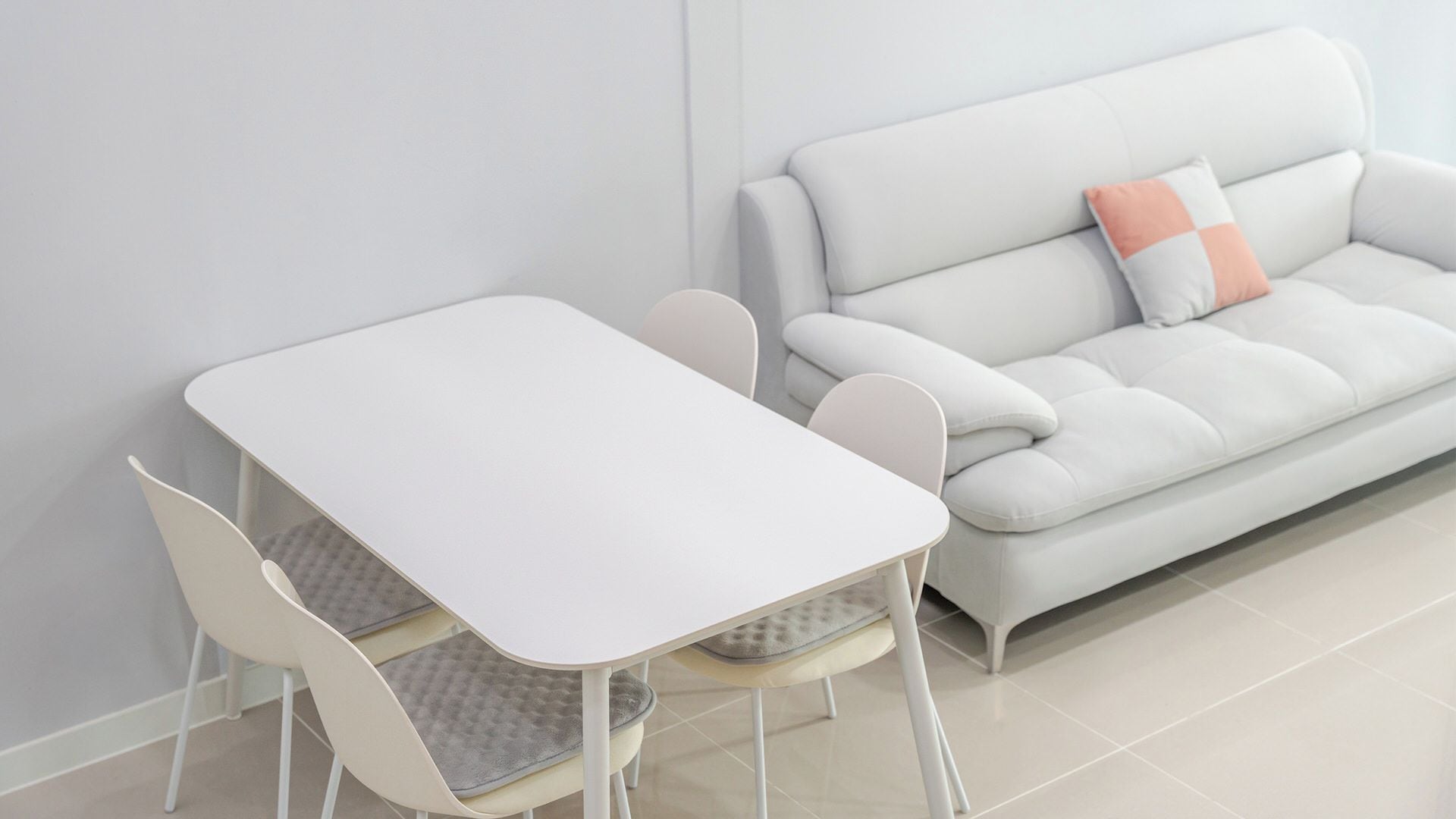 Habitació moderna i minimalista amb taula i cadires de color blanc a conjunt, un sofà gris amb un coixí sol i una paret blanca sense adorns.