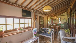 A room with a wooden ceiling, a fan, a sofa, a dining table, and a kitchen area. - Breeze and Blossom Cottage Maguwo (Depok)