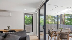 Una sala de estar moderna con un sofá gris, una mesa de comedor de madera y un amplio ventanal corredizo que da a un patio exterior con mobiliario de madera.