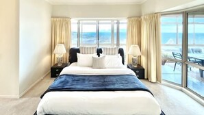 Ein Hotelzimmer mit einem großen Bett, zwei Nachttischen mit Lampen, Meerblick durch große Fenster und einem Balkon mit Sitzgelegenheit.