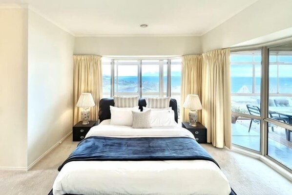 Quarto de hotel com uma cama grande, duas mesas de cabeceira com luminárias, vista para o mar por grandes janelas e varanda com mobiliário de exterior.