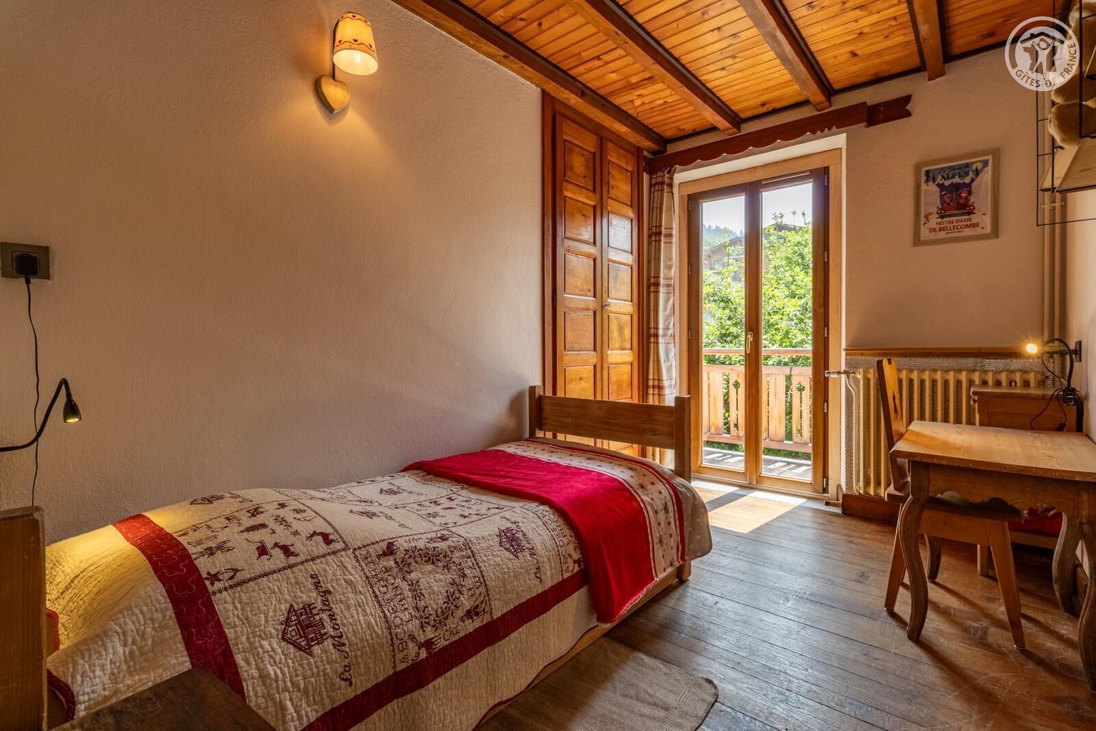Una habitación acogedora con cama, una mesa y una silla. Se ve follaje verde por la ventana.