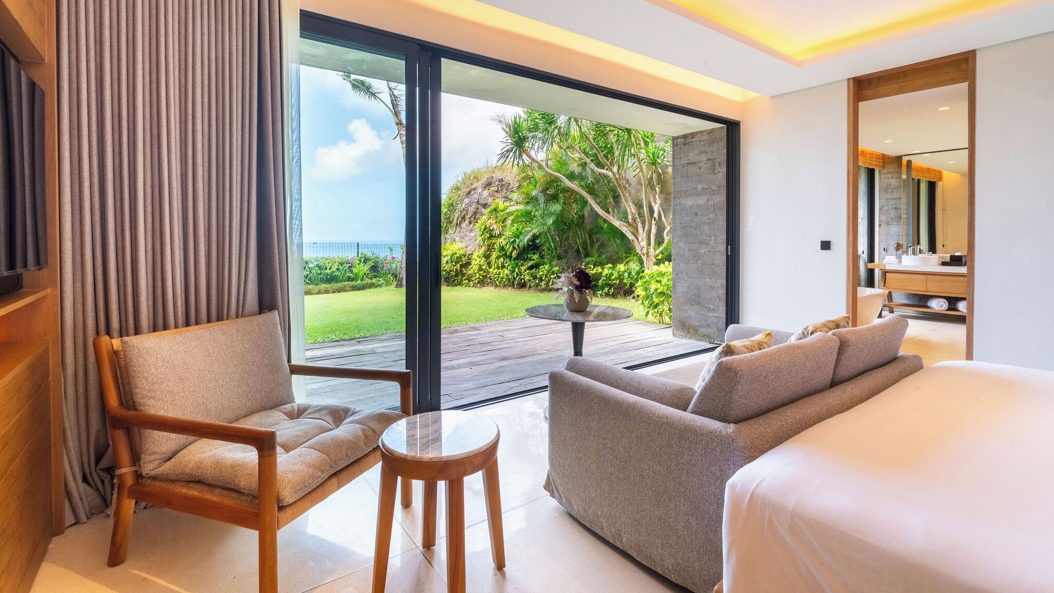 A modern hotel room with a large bed, a sofa, a chair, and a small table. There is a large window offering a view of a garden and the ocean.