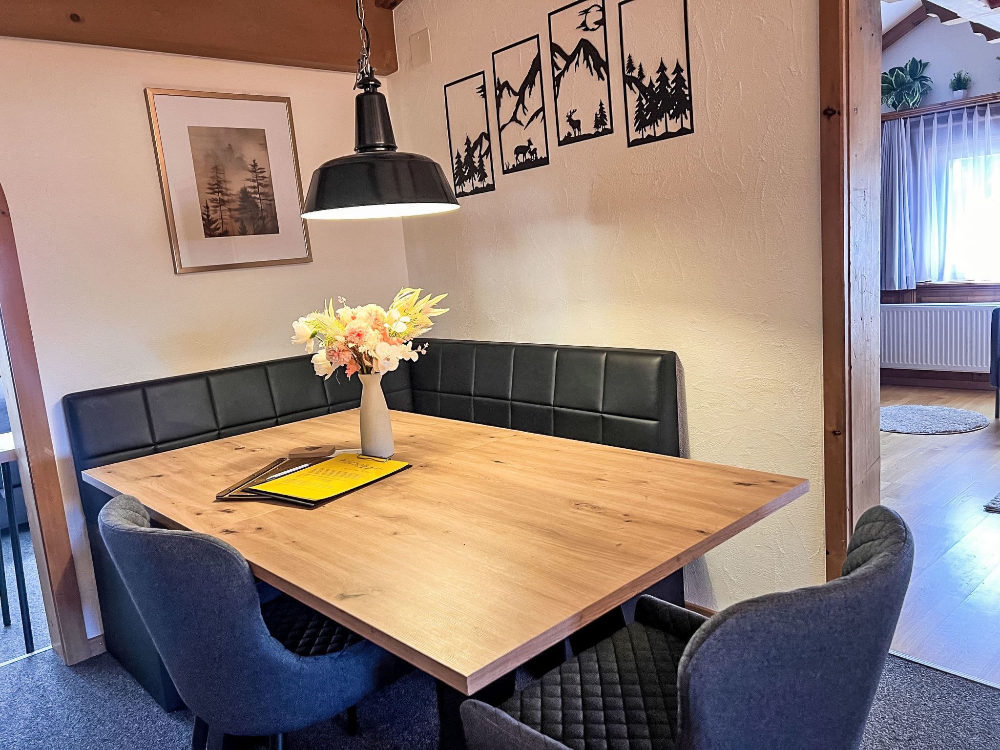 Un comedor con una mesa de madera, asientos tipo banca con cojines negros, un jarrón con flores, una lámpara colgante negra y obras de arte enmarcadas en la pared.