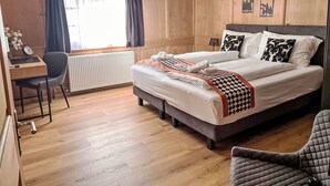 Habitación de hotel con pared de paneles de madera, cama con ropa blanca y almohadas estampadas, un escritorio con una silla y un radiador.