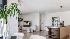 Un salon moderne avec une table  à manger en verre, des canapés blancs, un téléviseur et un îlot de cuisine doté d’un évier et d’étagères.