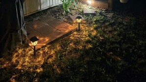 A house exterior at night with a wooden staircase, a small garden area with a pathway, and a porch light.