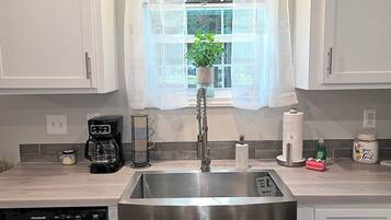 Una cocina con gabinetes blancos, un lavamanos de acero inoxidable, una cafetera y un dispensador de jabón para platos.