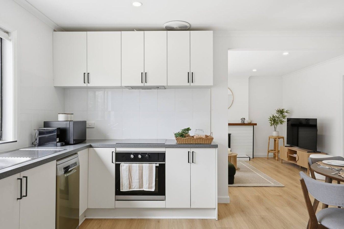 Una cocina moderna con armarios blancos, horno integrado y un comedor con televisor y asientos.
