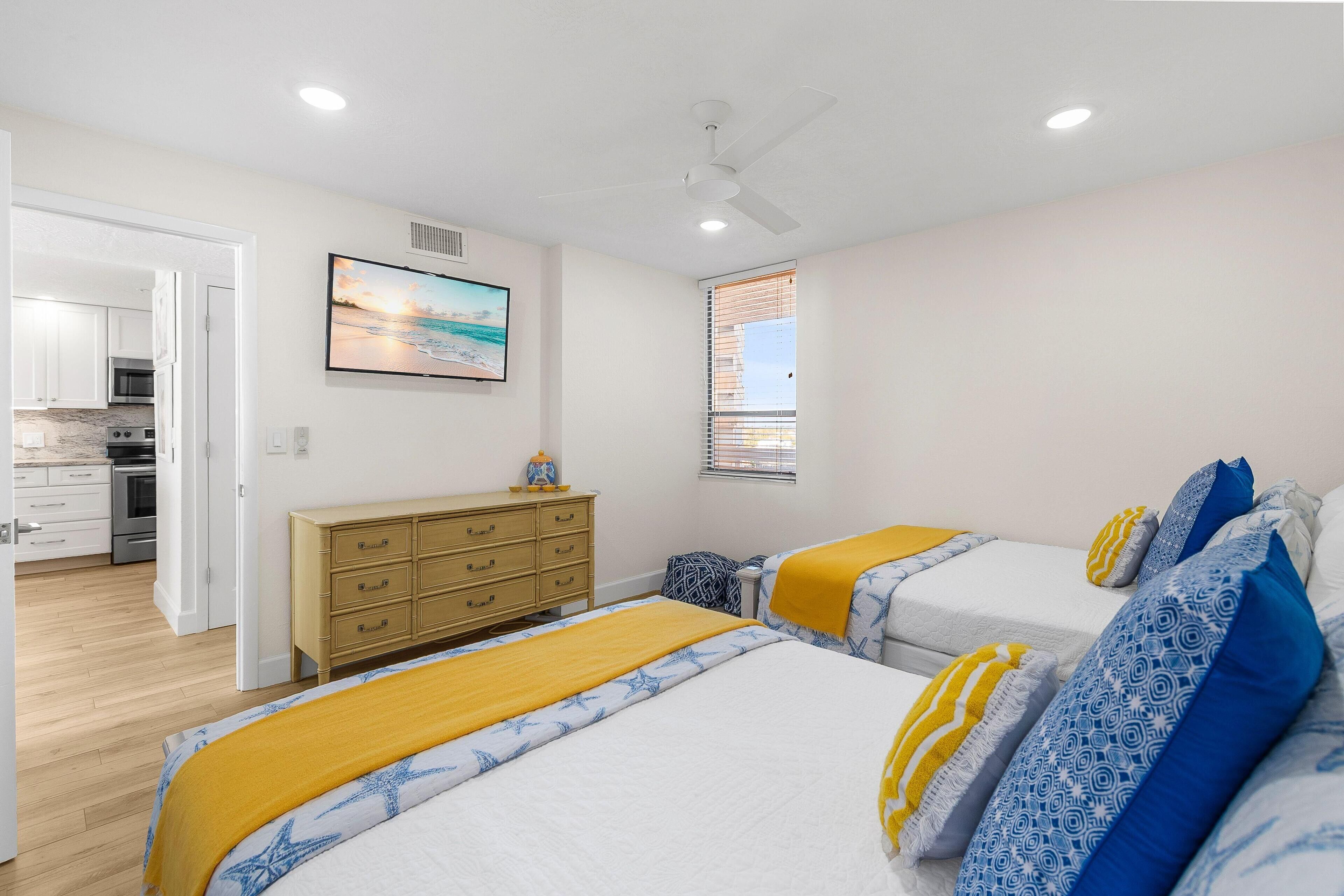 Une chambre avec deux lits, une commode, un ventilateur de plafond, un tableau et une fenêtre avec des stores.