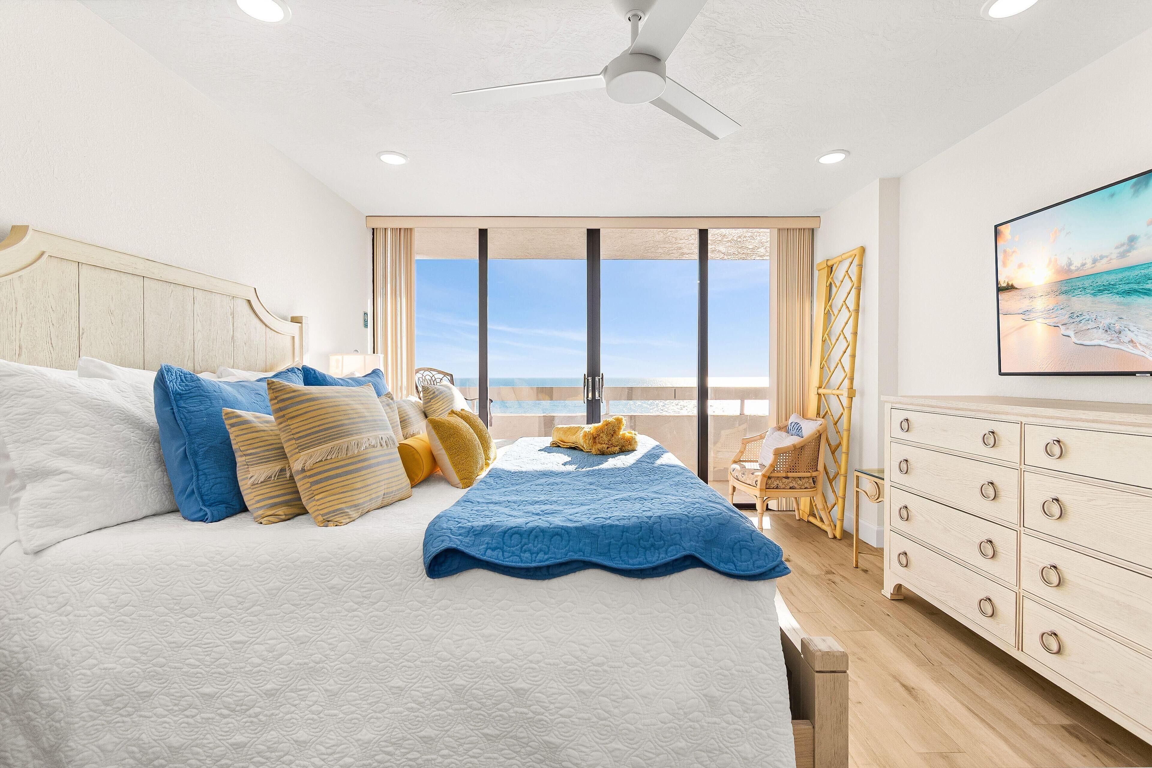 A bedroom with a large bed, a dresser, a ceiling fan, and a view of the beach.