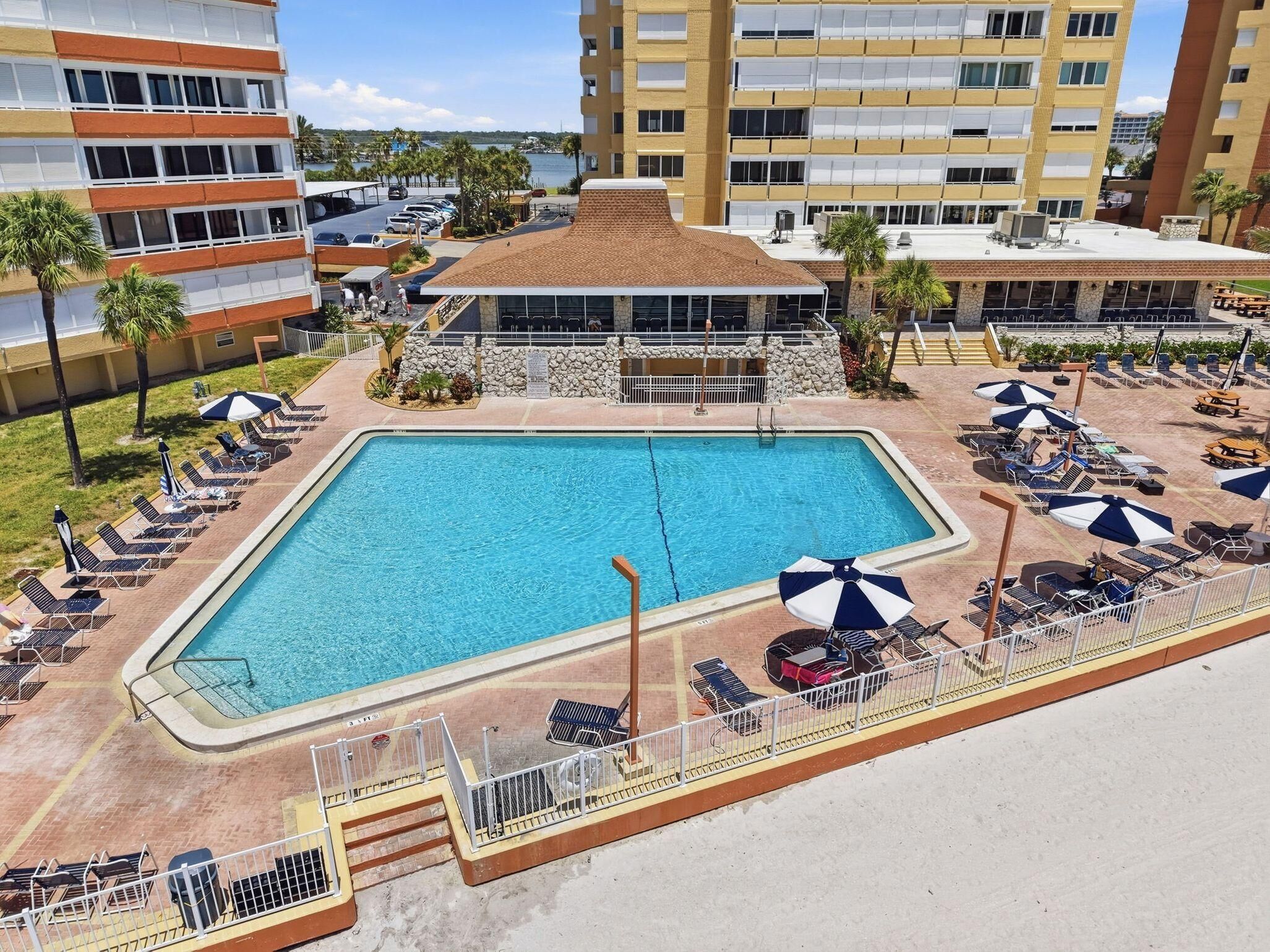 Ein rechteckiger Pool mit Liegestühlen und Sonnenschirmen, umgeben von einer Steinmauer und einem Gebäude mit mehreren Balkonen.