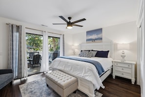 Une chambre à coucher avec un lit, des tables de chevet, un banc, un ventilateur de plafond et une vue sur l’extérieur, visible à travers une porte vitrée.