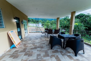 A covered outdoor area with a tiled floor, a table with chairs, and a view of greenery and buildings in the background. - Villa Verde - Scenic Views of El Yunque Rainforest (Río Grande)