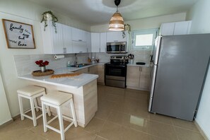 A modern kitchen with white cabinets, a stainless steel refrigerator, a microwave, and a gas stove. There are two white stools and a decorative sign on the wall. - Villa Verde - Scenic Views of El Yunque Rainforest (Río Grande)