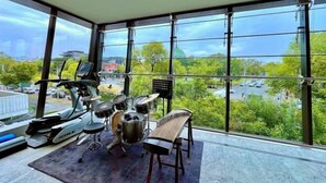 Quarto com uma janela grande, um conjunto de bateria, uma bicicleta ergométrica e um banco.