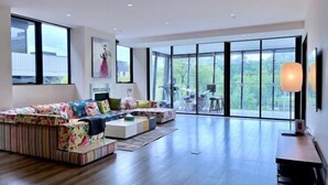 Uma sala de estar moderna com um sofá grande, uma mesa de centro e um espaço de jantar com cadeiras. Há uma janela ampla que oferece vista para o exterior.