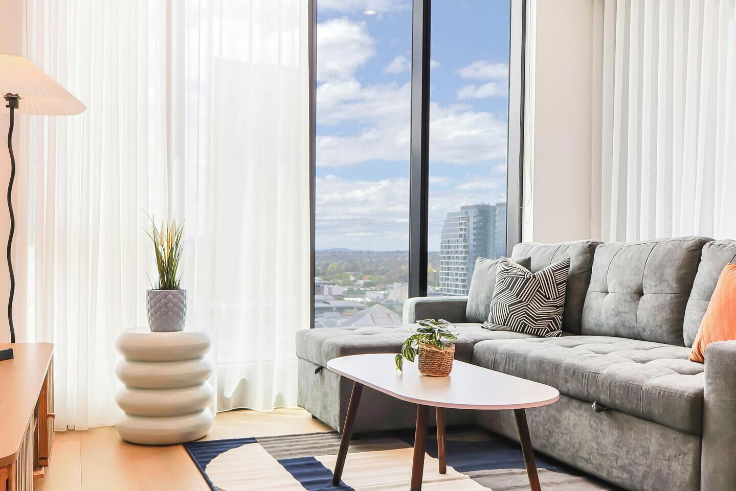 A modern living room with a grey sofa, a round coffee table, and a view of a cityscape through large windows.