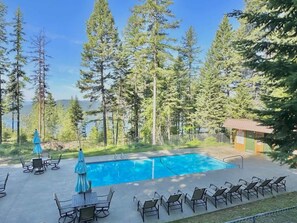 A rectangular swimming pool with lounge chairs and umbrellas, surrounded by tall trees and a small building. - Mountain Condo | Pool & Hot Tub | Perfect Location (Whitefish)