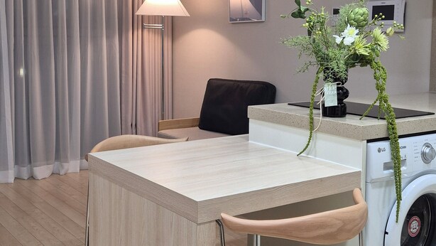 Een kleine keuken met een wasmachine, een eethoek met een tafel en stoelen, een lamp en een wandgemonteerde airconditioning.