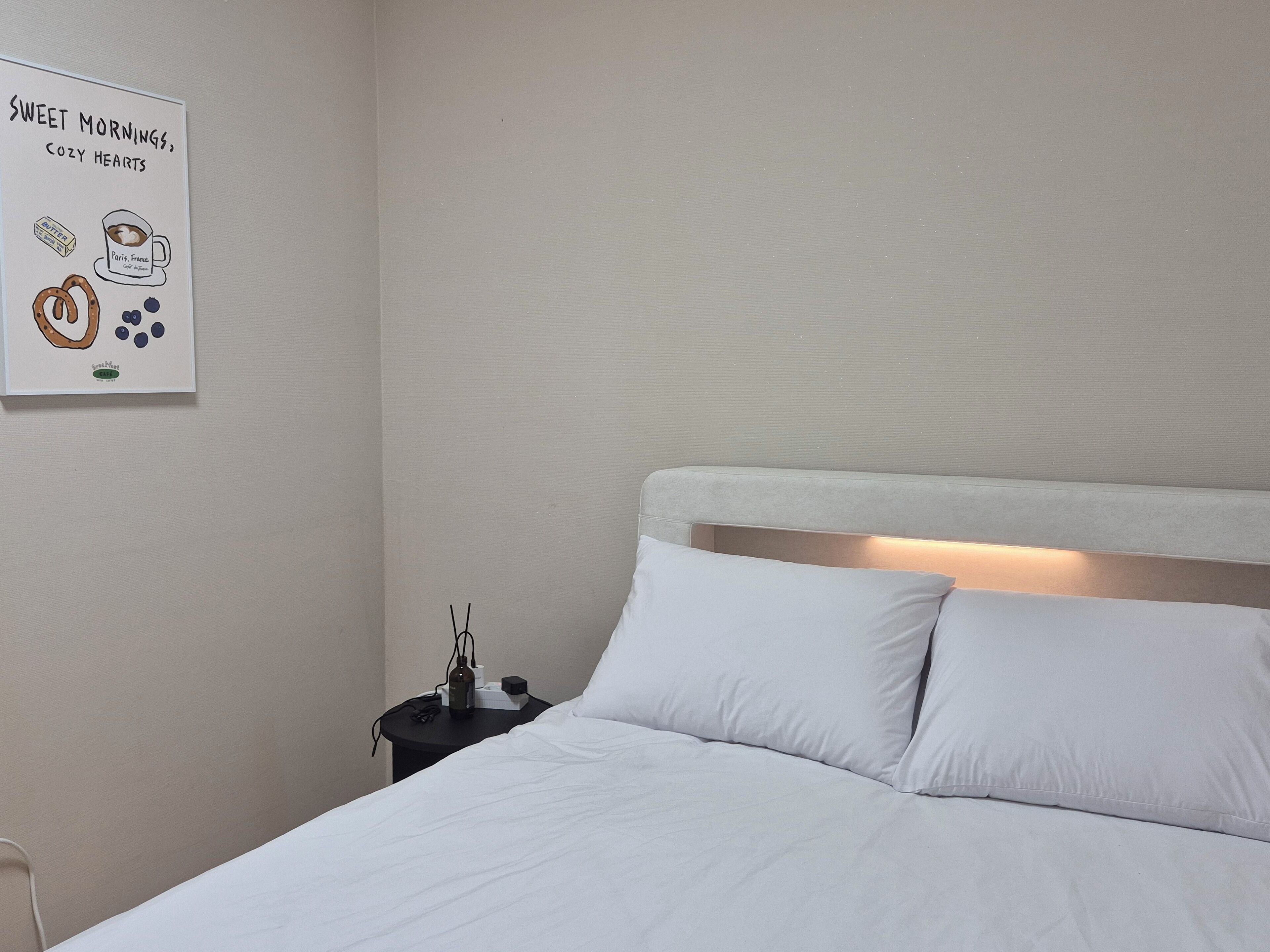 A neatly made bed with white linens, a bedside table with a lamp, and a wall-mounted sign with illustrations of a coffee cup, pretzel, and heart shapes.