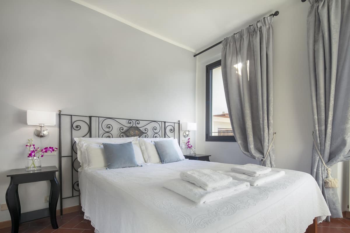 A neatly made bed with white linens, a bedside table with a lamp, and a window with curtains.