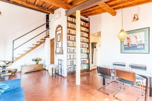 A spacious living area with a wooden staircase, a bookshelf, a dining table with chairs, and a large window. - Pitti Luxury Terrace (Florence)