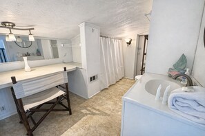 Un baño con tocador blanco, lavamanos, espejo, cortina de ducha y perchero.