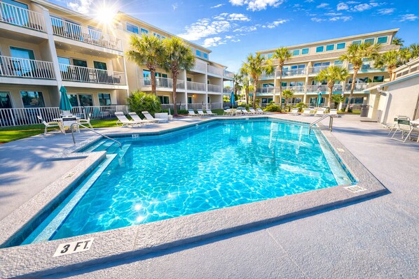 Una piscina rectangular con un indicador de profundidad de 3 pies, rodeada de sillas de descanso y palmeras, con un edificio de varios pisos al fondo.