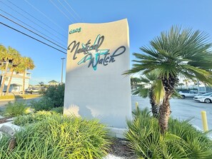 Ein hohes, beiges Schild mit den Wörtern 'Nautical' und 'Beach' in stilisierter Schrift, umgeben von Grünpflanzen und Palmen.