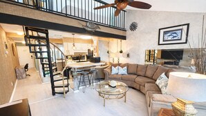 Ein großzügiger Wohnbereich mit einer großen Couch, einem Couchtisch und einem Fernseher. Eine Treppe führt zu einer oberen Ebene.