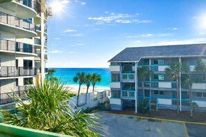 Un hotel frente a la playa con varios balcones con vista al océano, un estacionamiento y palmeras.