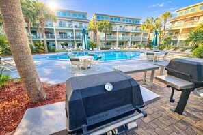 Zona de piscina con sillones reclinables, una parrilla y un edificio con balcones al fondo.