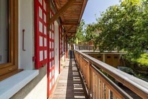 Un balcón de madera con contraventanas rojas, barandilla de madera y vistas a zonas verdes y edificios al fondo.