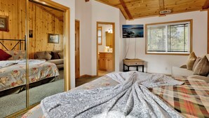 Een gezellige kamer met houten wanden en plafond, een bed met bloemenbedrukking, een bank, een kleine tafel en een raam met jaloezieën.