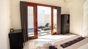 A bedroom with a bed, a desk, a wardrobe, and a view of an outdoor pool area. - Villa Taman Bali 6 (Kerobokan)
