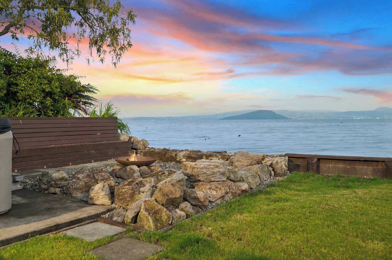 A stone fire pit with a bench and a wooden deck overlooking a body of water with distant landforms.