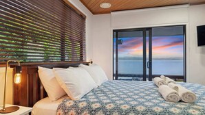 Une chambre à coucher avec un grand lit, une table de chevet, une lampe et une vue sur la mer, visible à travers une porte vitrée.