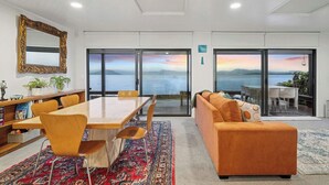 Un salon moderne avec une grande table à manger en bois, un canapé orange et une vue sur la mer grâce à de grandes portes coulissantes.