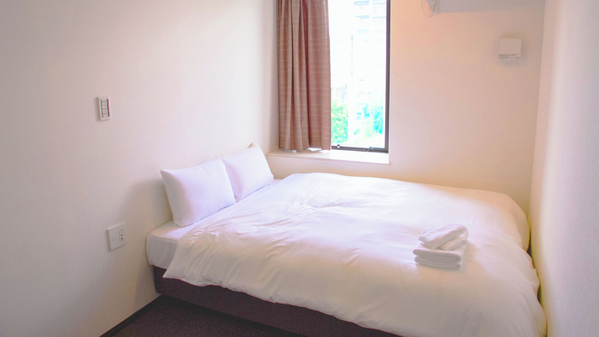 Ein Einzelbett mit weißer Bettwäsche, ein Fenster mit Vorhängen und ein Thermostat an der Wand.