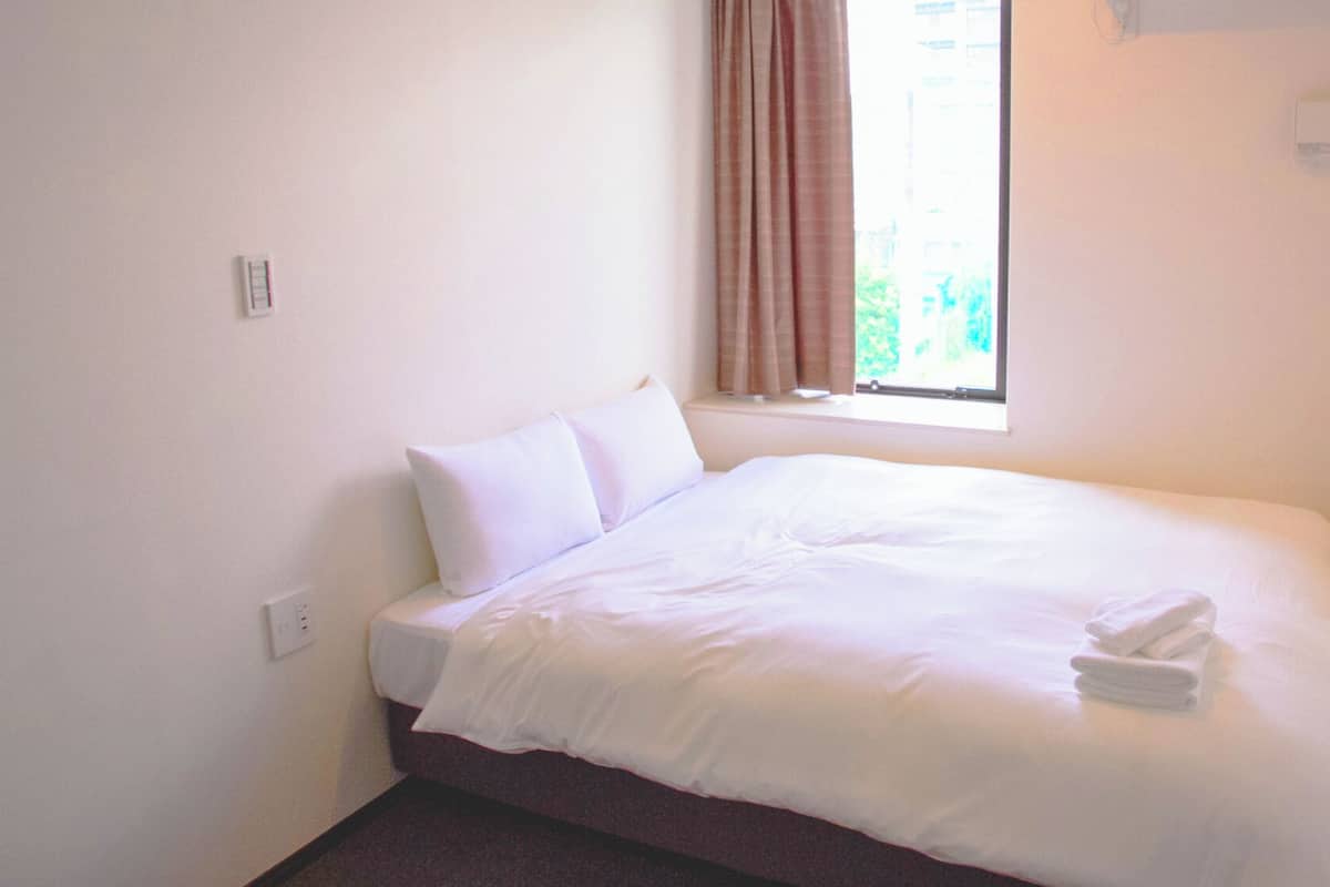 Ein Einzelbett mit weißer Bettwäsche, ein Fenster mit Vorhängen und ein Thermostat an der Wand.