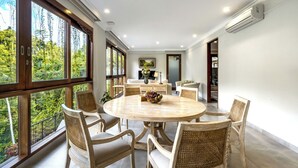Un coin repas avec une table ronde, six chaises en osier et une vue sur la verdure grâce à de grandes fenêtres.