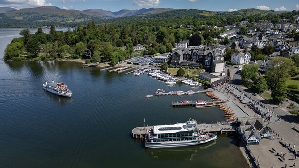 Une vue aérienne d’une ville au bord d’un lac, avec un grand quai, plusieurs bateaux et un bâtiment imposant surmonté d’un clocher.