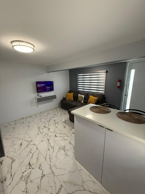 A modern living room with a flat-screen TV, a sofa with cushions, a window with blinds, and a kitchen counter with woven placemats. - Cozy, centrally located, family-friendly apt (Cataño)