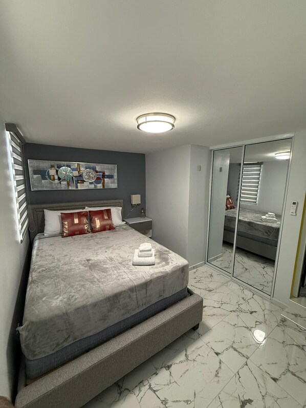Une chambre moderne avec un grand lit, une table de chevet, une commode, une œuvre d’art accrochée au mur et un dressing avec une porte de placard miroir.