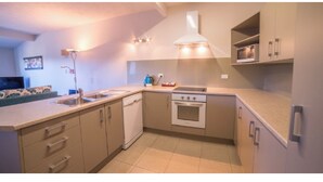 A compact kitchen with a dishwasher, oven, and microwave. There is a sink with a faucet, and cabinets with handles. A ceiling light fixture is visible above the sink. - Paihia resort with all the amenities! (Paihia)