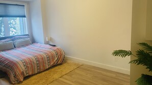 A bedroom with a bed, a window with blinds, a nightstand, a lamp, a fluffy rug, wooden flooring, and a potted plant. - Stunning 3 bedroom modern apartment in Downtown Toronto (Toronto)