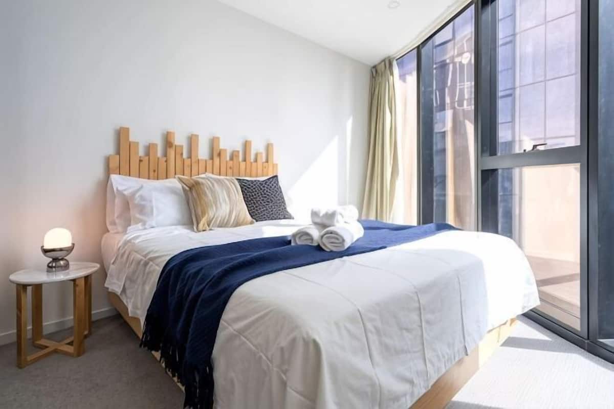 Een moderne hotelkamer met een groot bed, een nachtkastje met een lamp en uitzicht door een groot raam.