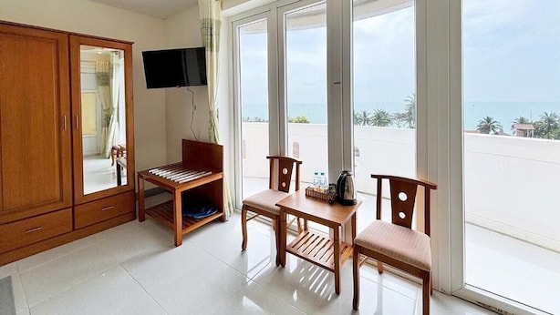 A room with a wooden cabinet, a television, a table with chairs, and large windows offering a view of the sea.