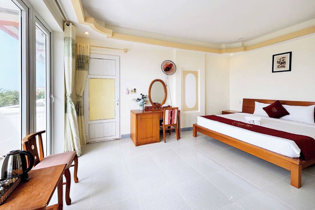 Une chambre d’hôtel comprenant un lit, une commode en bois, un bureau avec une chaise, un ventilateur et une fenêtre avec des rideaux.
